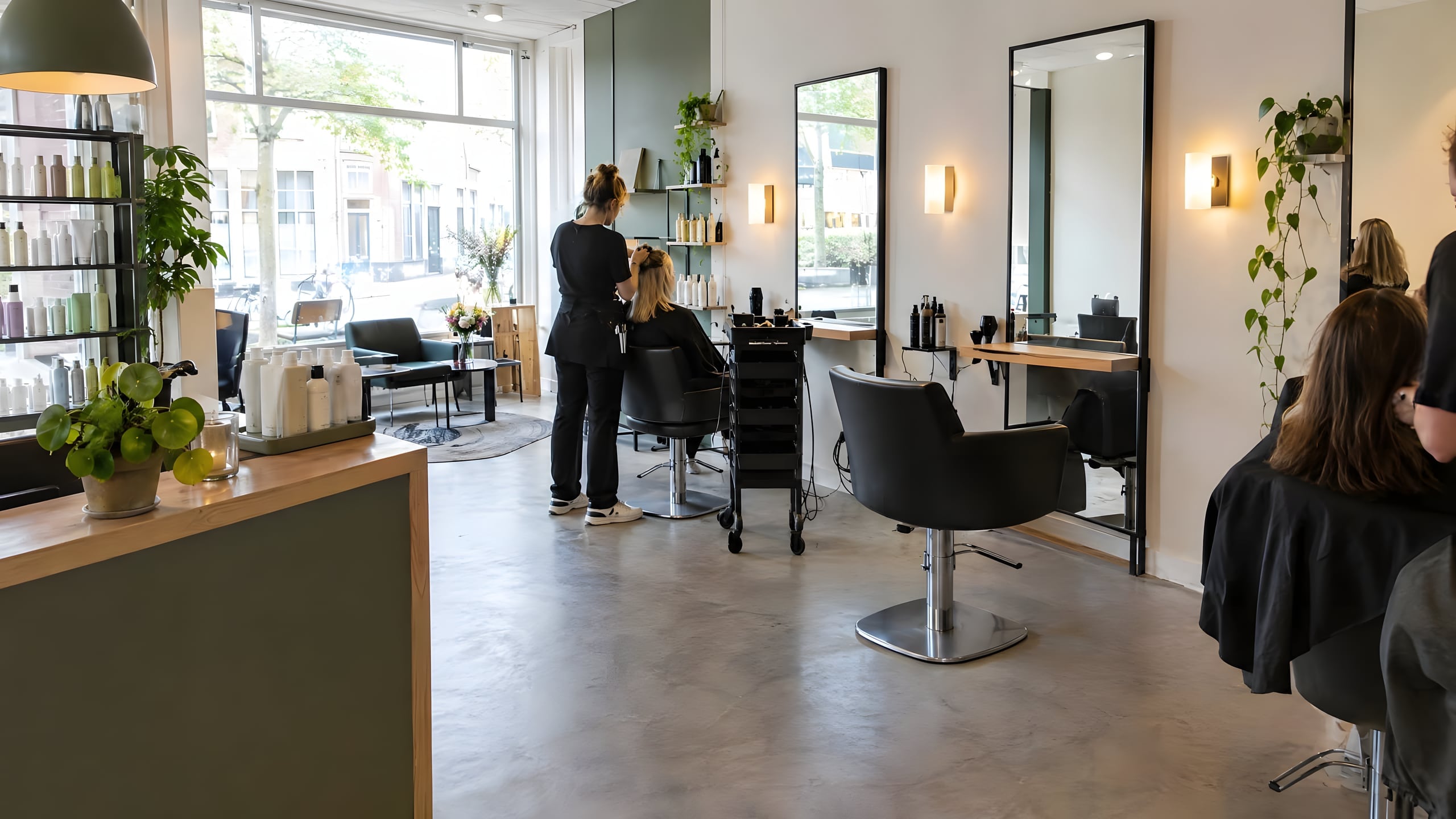 Het interieur van een lichte kapsalon met spiegels, kappersstoelen en planten bij het raam.
